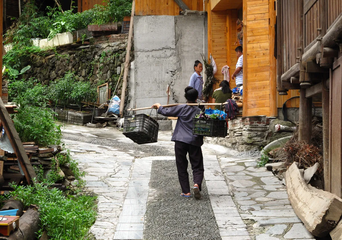 木造家屋が並ぶ石畳の路地を、天秤棒でかごを運びながら歩く人物の後ろ姿