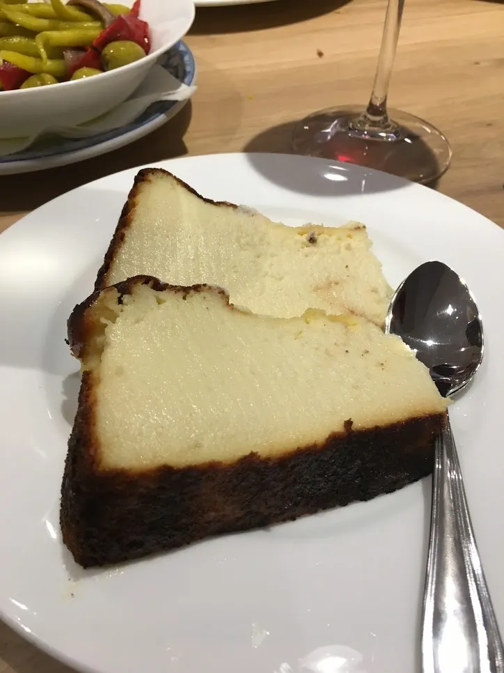 白い皿に置かれた三角形の一切れのケーキ。上面と側面全体が濃い焦げ茶色から黒に変色し、切り口はクリーム色でつやがあり、中心に近いほど柔らかそうな質感。右にスプーン、奥にグラスと小鉢