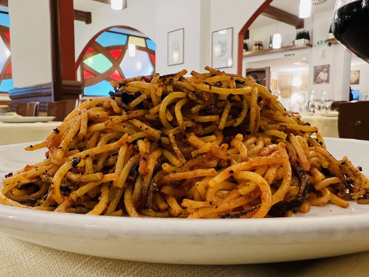 白い深皿に、赤茶色に色づいた太めのスパゲッティが山盛りになっている。麺の縁のあちこちに黒く焦げた部分が見える。背景は店内の柱とステンドグラス。
