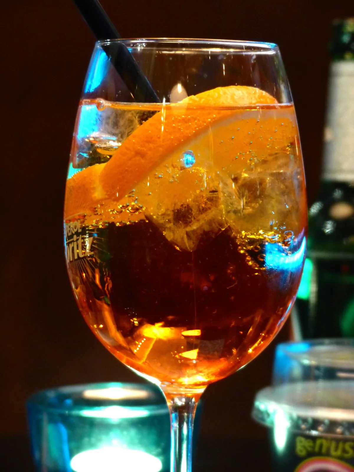 脚付きのワイングラスにオレンジ色の炭酸飲料が満たされ、オレンジの薄切りが縁に掛かる。氷の間から細かい気泡が立ち上り、黒いストローが挿さる。グラス越しに「Aperol Spritz」と書かれたコースターが透けて見える。背景は暗く、青緑色の照明が反射