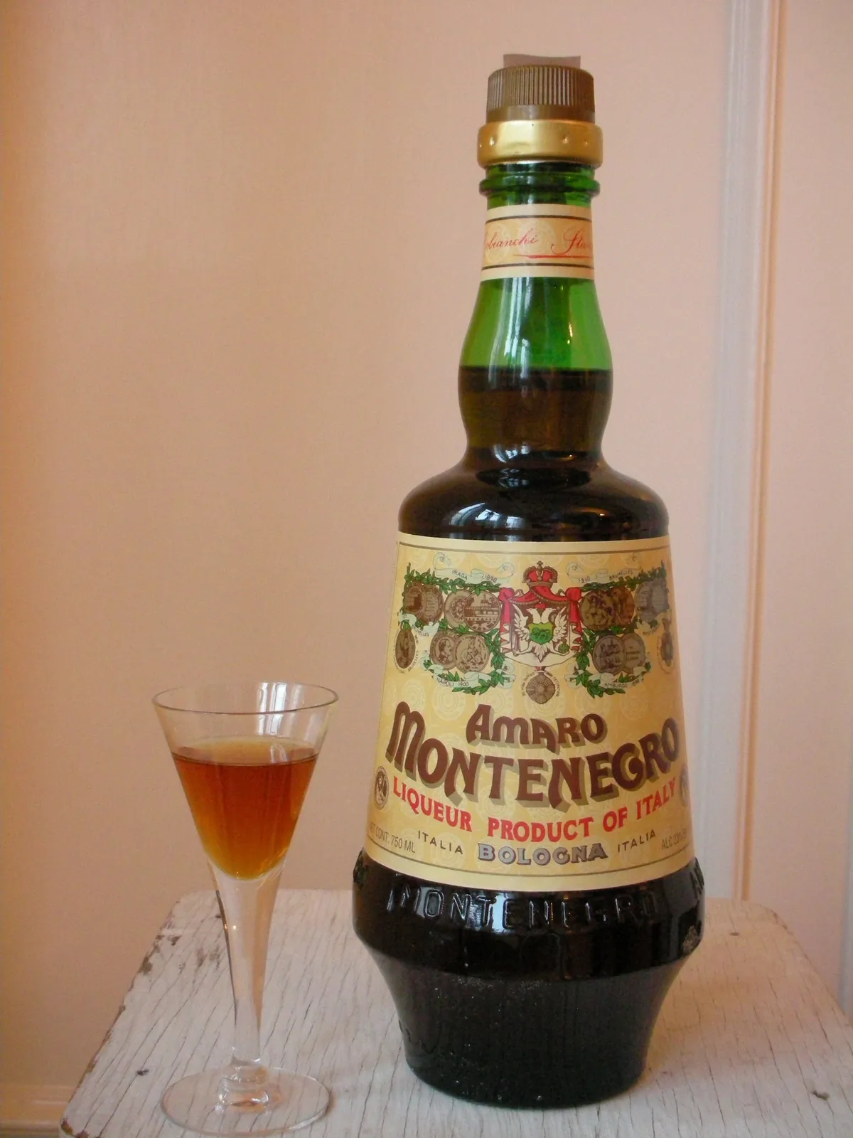 緑色の首をした濃茶色の瓶に、クリーム色のラベルで「AMARO MONTENEGRO」「LIQUEUR PRODUCT OF ITALY」「BOLOGNA ITALIA」と書かれている。その左に脚付きの小さなグラスが置かれ、琥珀色の液体が注がれている