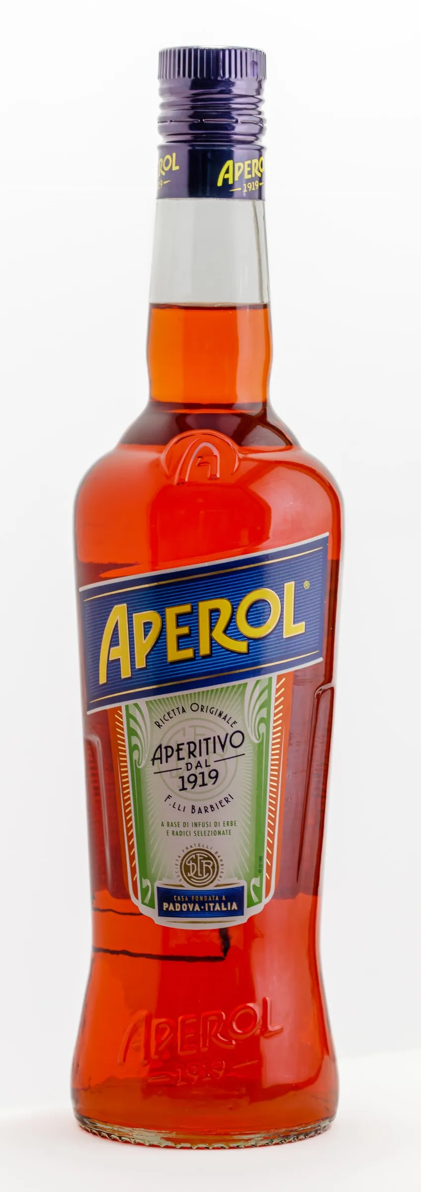 オレンジ色の液体が満たされたガラス瓶に、濃紺の帯に黄色で「APEROL」と書かれたラベルが中央に貼られ、その下に緑と白の帯で「Aperitivo dal 1919」の文字がある。背景は無地の白