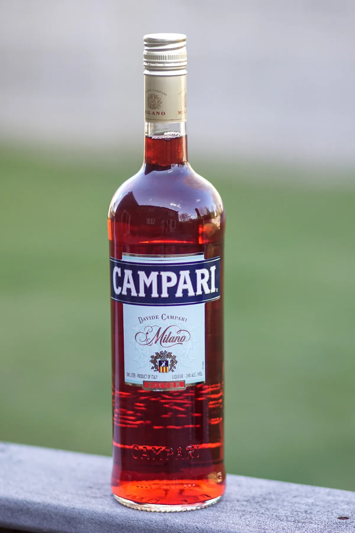 透明なガラス瓶に赤い液体が満たされ、紺色の帯に白抜きで「CAMPARI」と書かれた長方形のラベルが中央に貼られている。背景はぼけた緑の芝生と灰色の空