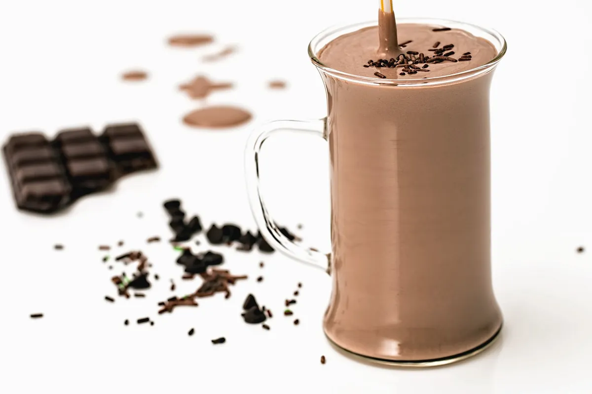 取っ手付きの透明なガラスマグに、薄茶色のチョコレートミルクがなみなみと注がれ、表面に細かい黒いチョコレートの粒が散っている。手前には板チョコレートの欠片が置かれている。