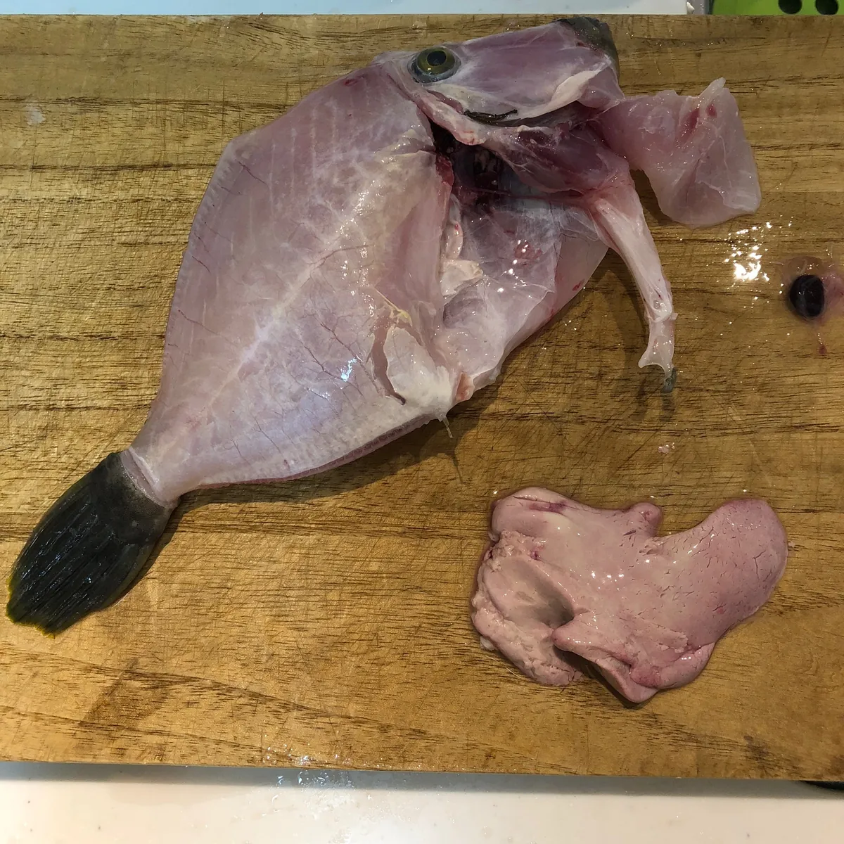木製のまな板の左側に、皮を剥かれ白っぽいピンク色をした魚体（目と尾が確認できる）、右側に血管の筋が透ける淡いピンク色で不定形の肝臓が置かれている。上部には胆嚢とみられる黒い粒が付着