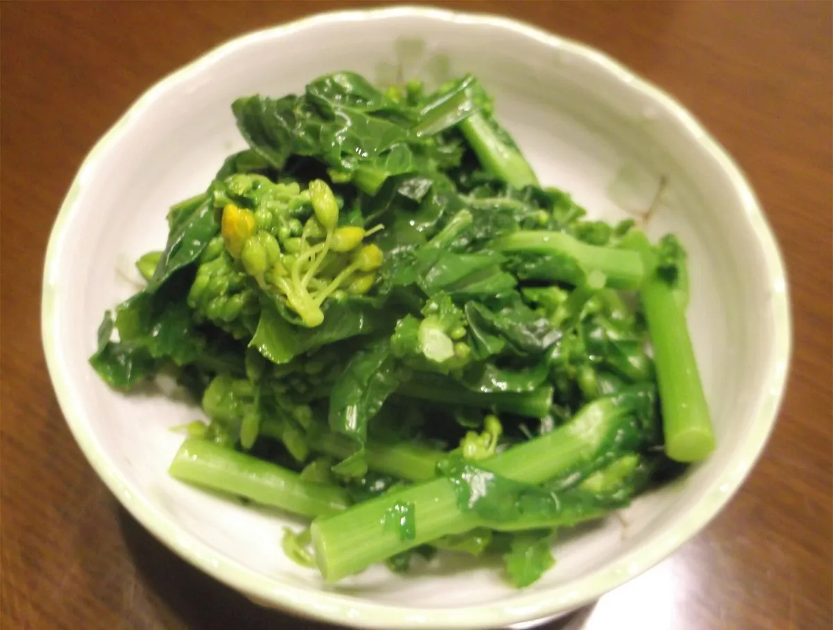 白い器の中に、濃い緑色に茹で上がった葉と茎が折り重なり、その一部に黄色い花芽の塊が覗いている。表面はつゆで軽く濡れて艶がある。