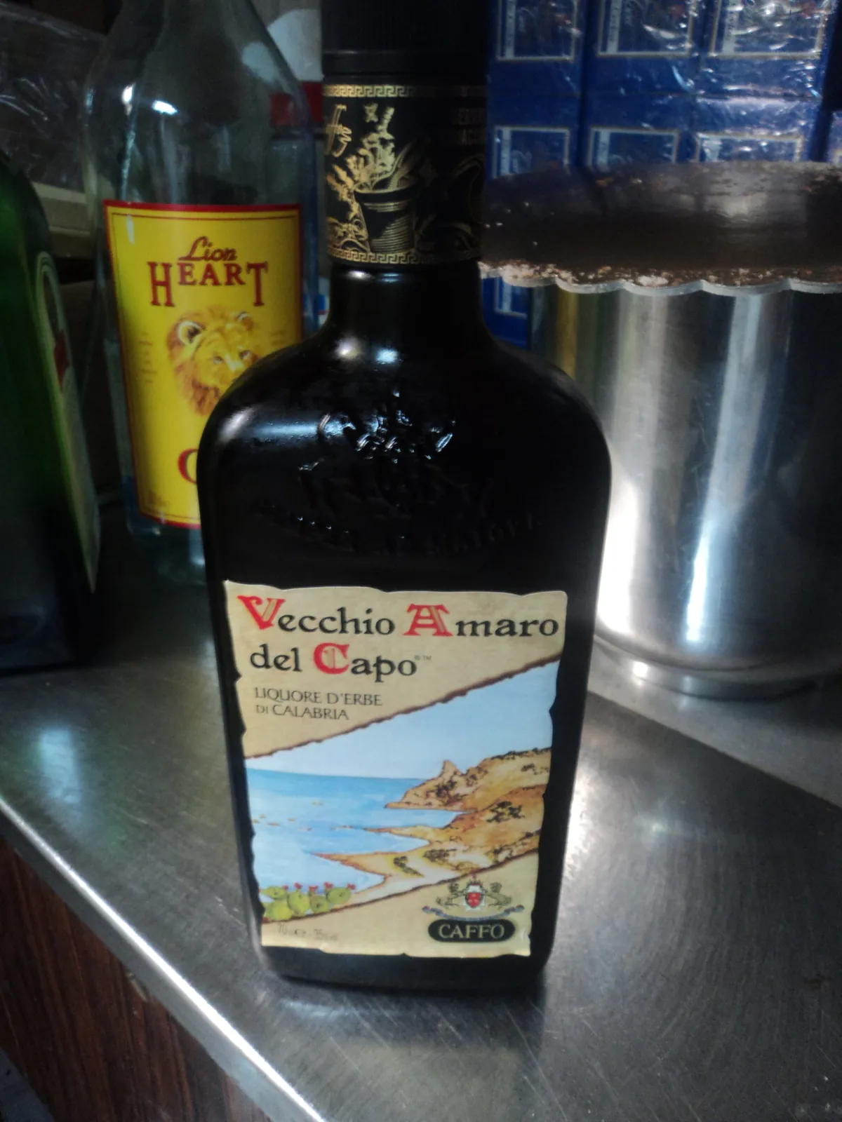 濃い茶色のずんぐりした瓶に、クリーム色の台形ラベルで「Vecchio Amaro del Capo」「Liquore d’Erbe di Calabria」「CAFFO」と書かれ、海岸線のイラストが描かれている。背景には別のボトルや金属缶が並ぶ棚