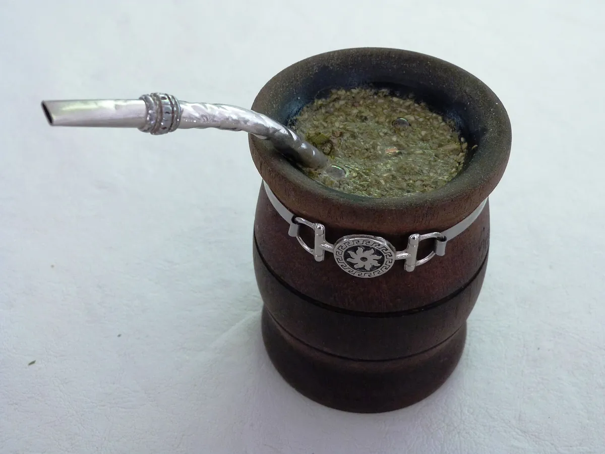 白い背景の上に置かれた樽形の木製カップの中に、湿った緑褐色の茶葉が縁近くまで詰まっている。カップの縁からは装飾の入った銀色の曲がったストローが差し込まれている。