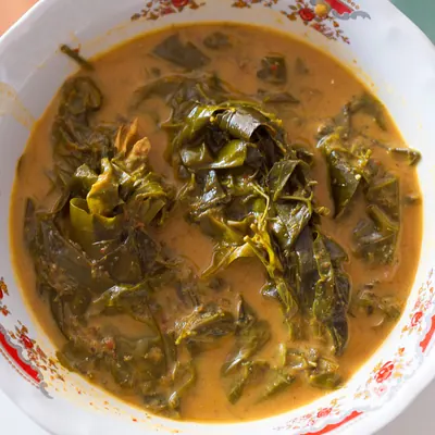 花柄の縁取りがある白い器に、赤みを帯びた黄褐色のとろみあるカレースープが満ち、しんなりと煮えた濃緑色の葉が塊になって浮かんでいる真上からの写真。