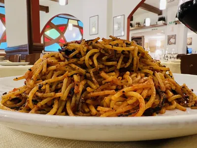 白い深皿に、赤茶色に色づいた太めのスパゲッティが山盛りになっている。麺の縁のあちこちに黒く焦げた部分が見える。背景は店内の柱とステンドグラス。