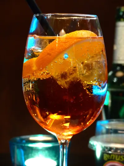 脚付きのワイングラスにオレンジ色の炭酸飲料が満たされ、オレンジの薄切りが縁に掛かる。氷の間から細かい気泡が立ち上り、黒いストローが挿さる。グラス越しに「Aperol Spritz」と書かれたコースターが透けて見える。背景は暗く、青緑色の照明が反射