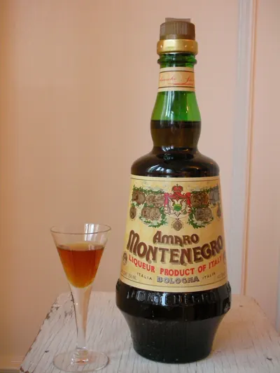 緑色の首をした濃茶色の瓶に、クリーム色のラベルで「AMARO MONTENEGRO」「LIQUEUR PRODUCT OF ITALY」「BOLOGNA ITALIA」と書かれている。その左に脚付きの小さなグラスが置かれ、琥珀色の液体が注がれている