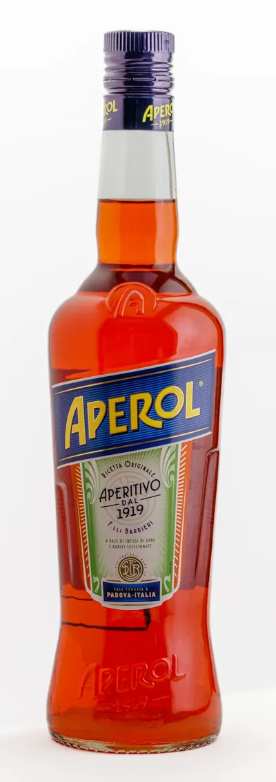 オレンジ色の液体が満たされたガラス瓶に、濃紺の帯に黄色で「APEROL」と書かれたラベルが中央に貼られ、その下に緑と白の帯で「Aperitivo dal 1919」の文字がある。背景は無地の白