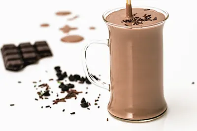 取っ手付きの透明なガラスマグに、薄茶色のチョコレートミルクがなみなみと注がれ、表面に細かい黒いチョコレートの粒が散っている。手前には板チョコレートの欠片が置かれている。